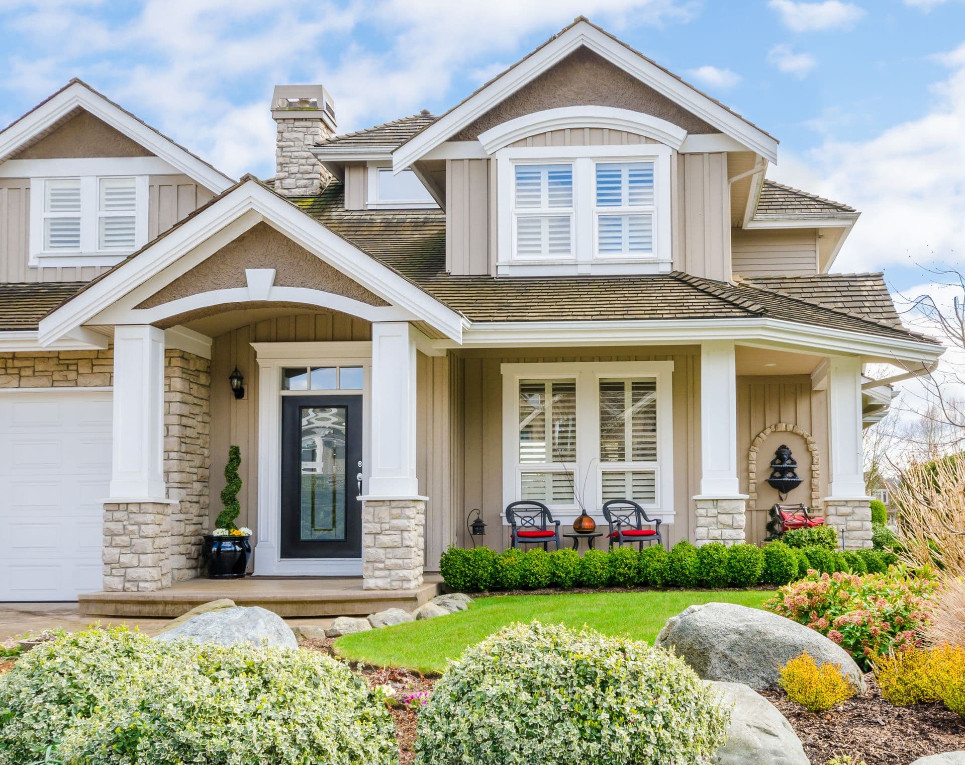 Charming suburban house with stone and beige siding, white trim, and gabled roof. Front porch has red chairs, greenery, and a lantern. Inviting and cozy atmosphere.