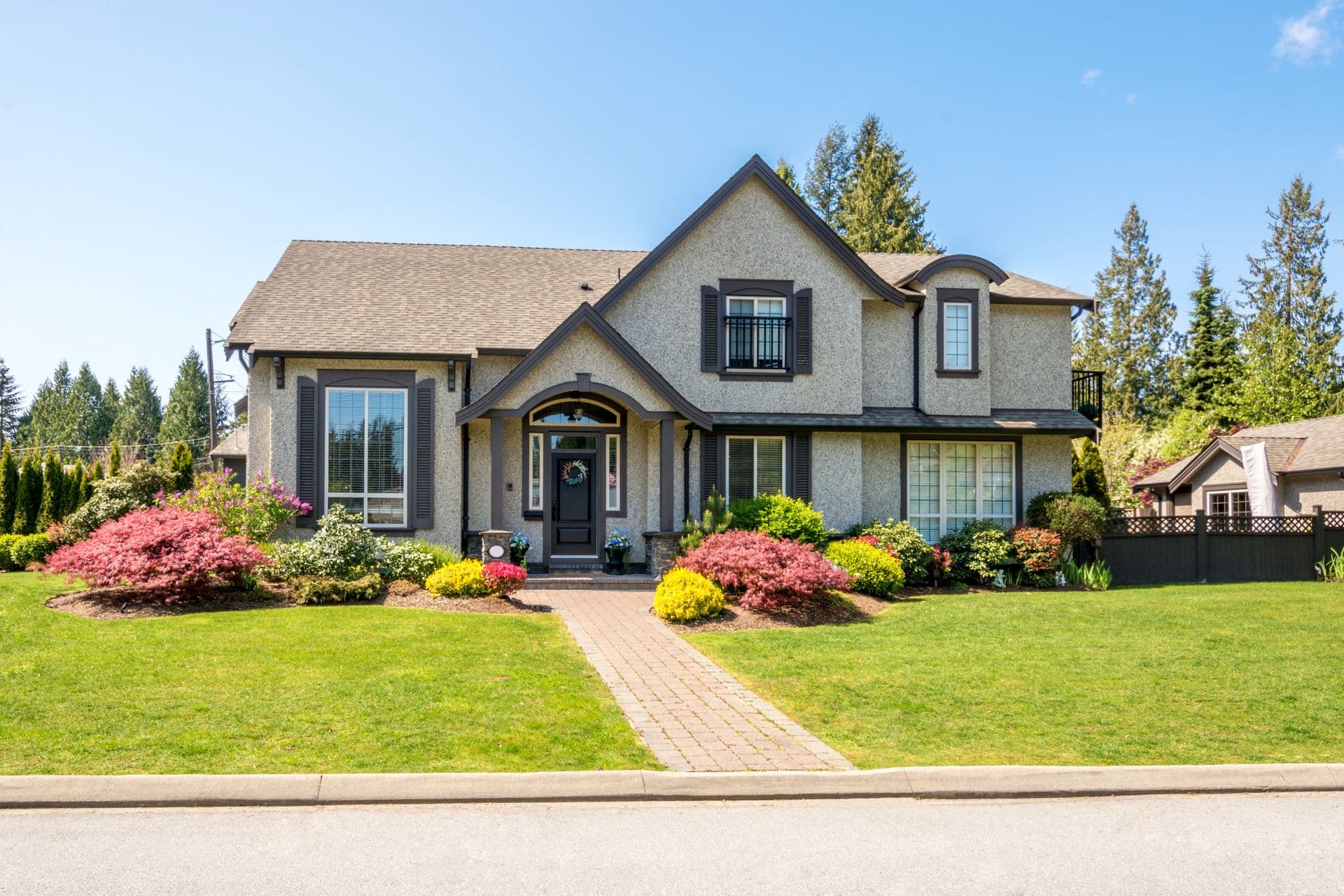 Charming two-story suburban house with gray stucco, dark shutters, and a manicured lawn. Vibrant flower bushes enhance the welcoming atmosphere.
