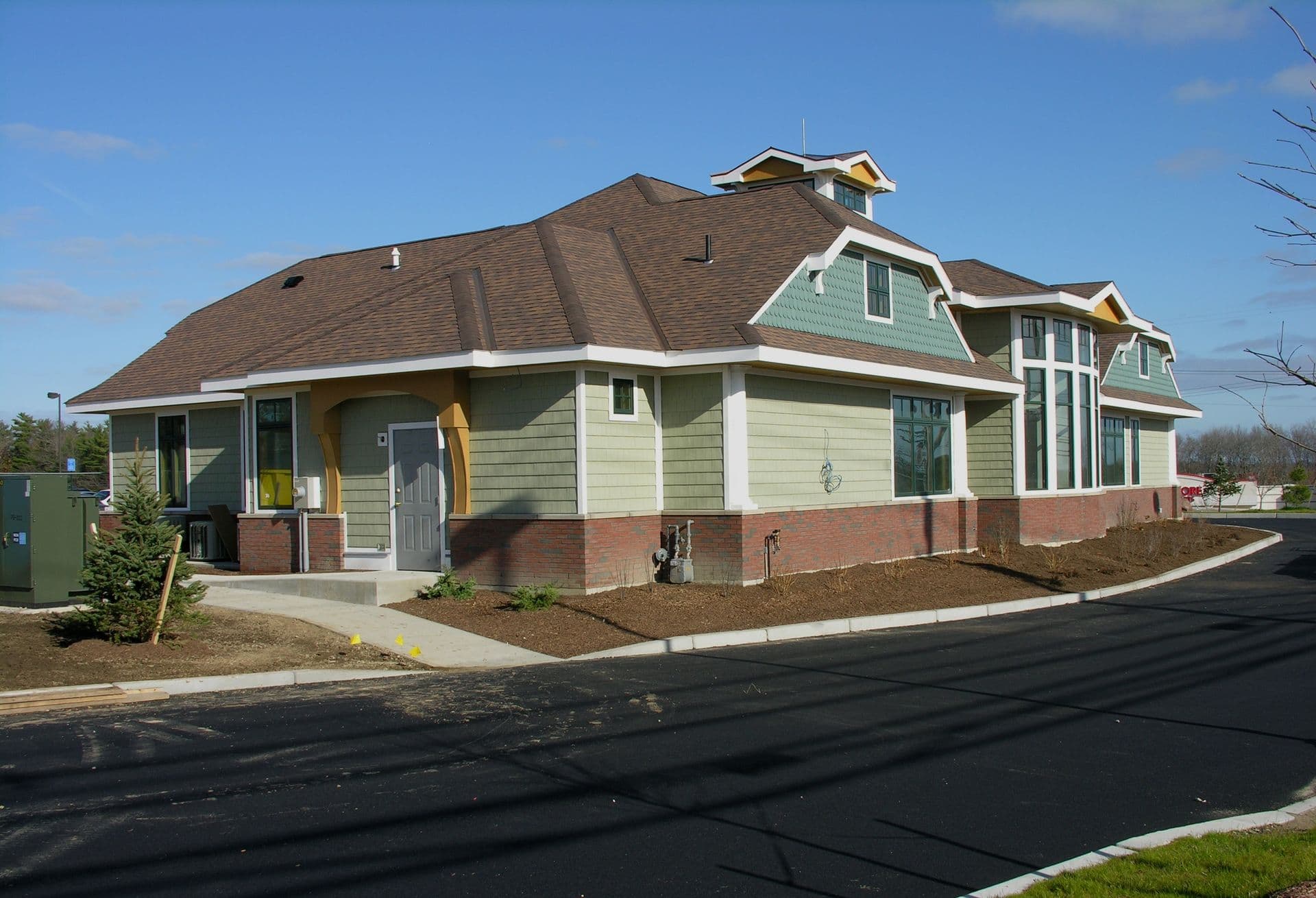 A modern, single-story building featuring green siding and brown gabled roof. Large windows and a fresh landscape contribute to a welcoming appearance.