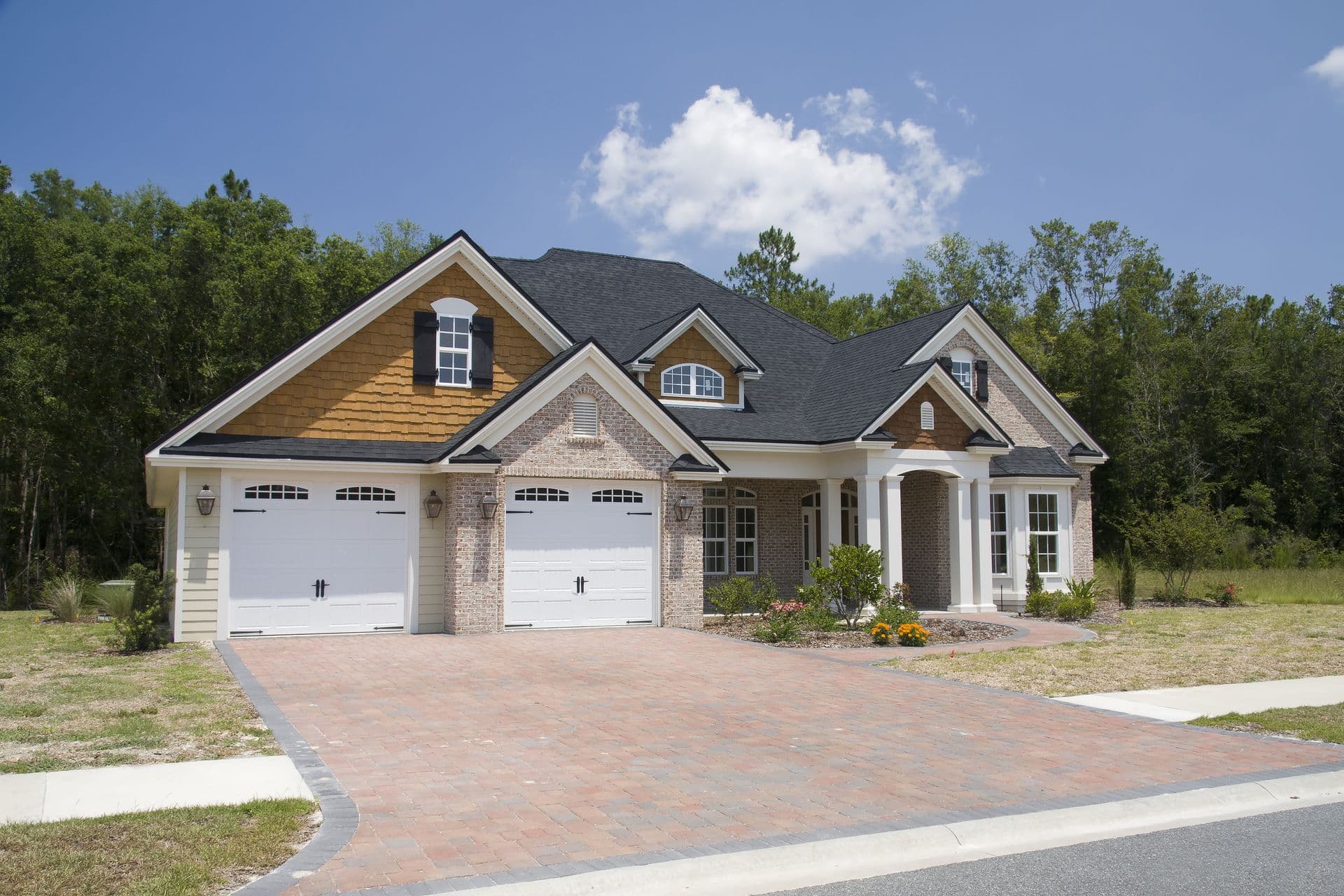 Single-story brick house with black roof, two white garage doors, and a paved driveway. Surrounded by greenery, under a clear blue sky.