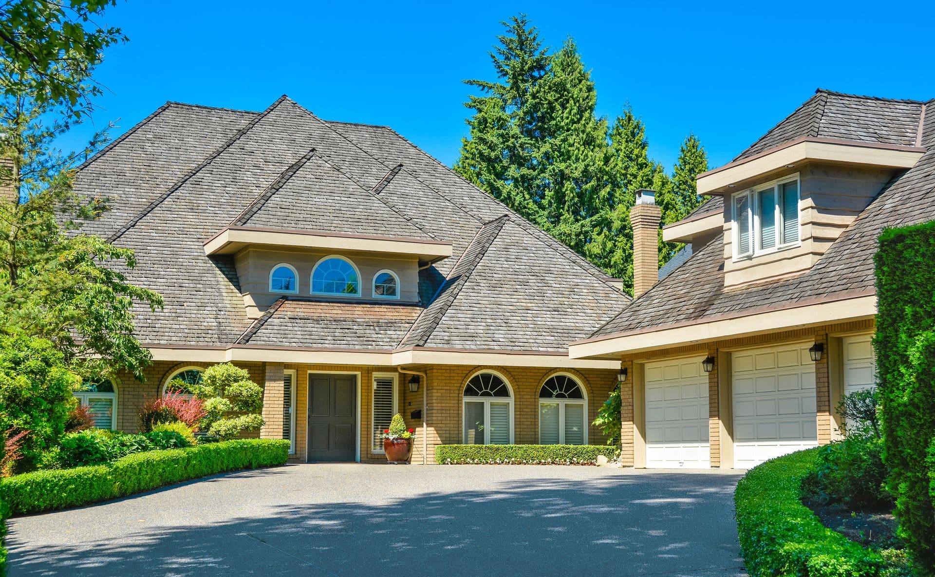 Luxurious brick house with triangular rooftops, large arched windows, and a double garage. Surrounded by green trees and hedges under a clear blue sky.