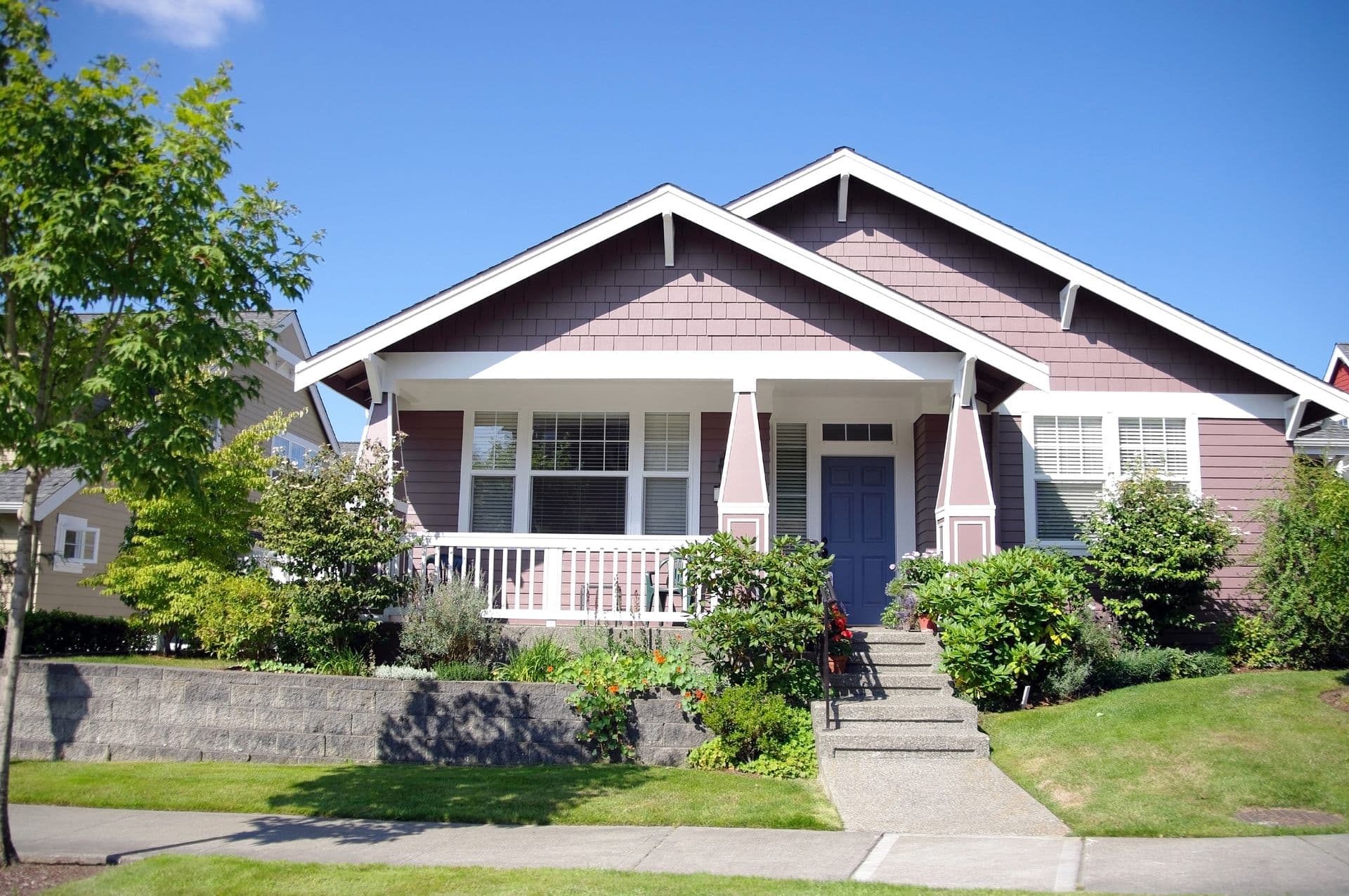 Charming purple suburban house with a white porch, surrounded by lush greenery and a manicured lawn, under a clear blue sky.