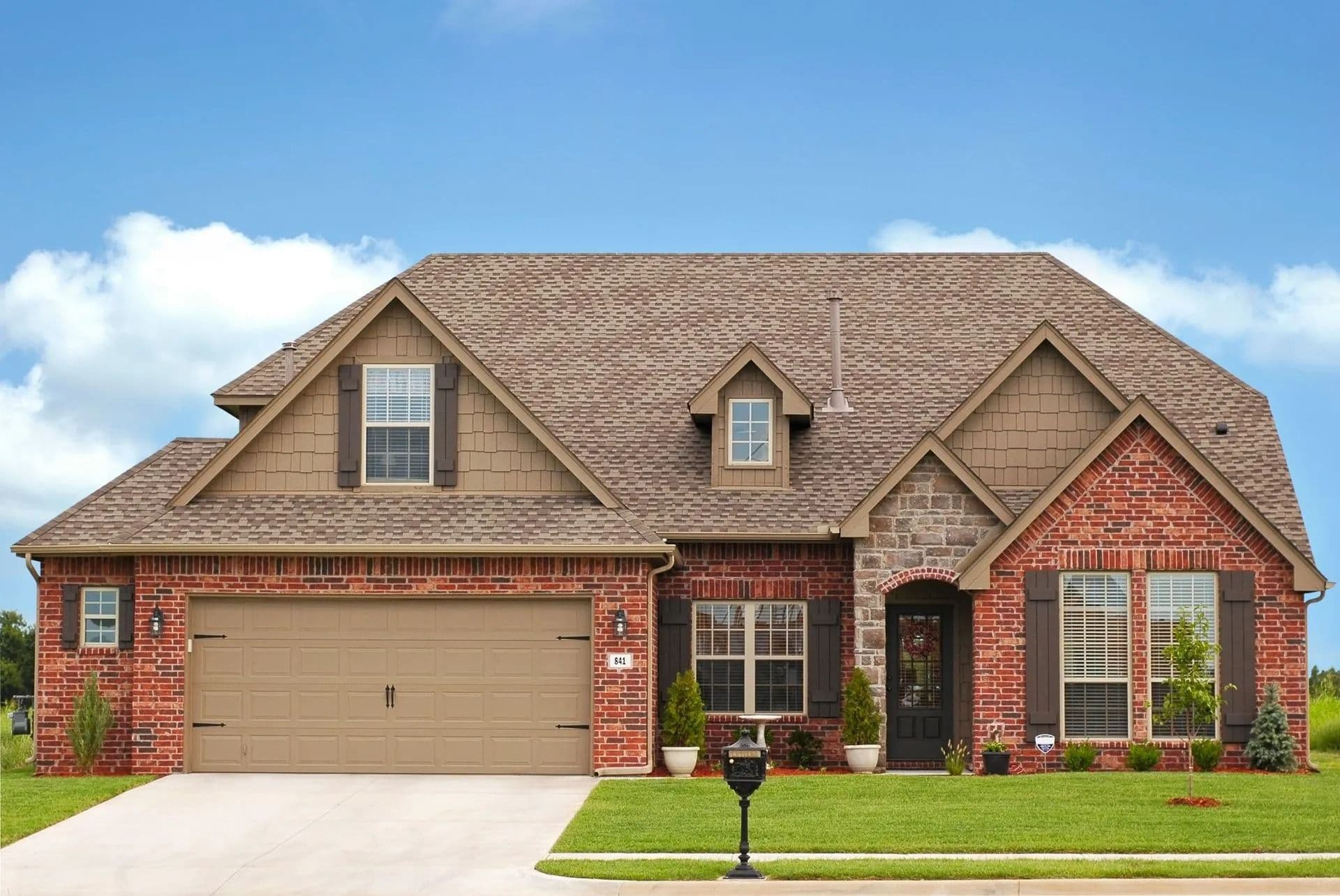 A red brick suburban house with a two-car garage and manicured lawn under a bright blue sky. The front features large windows and decorative plants.